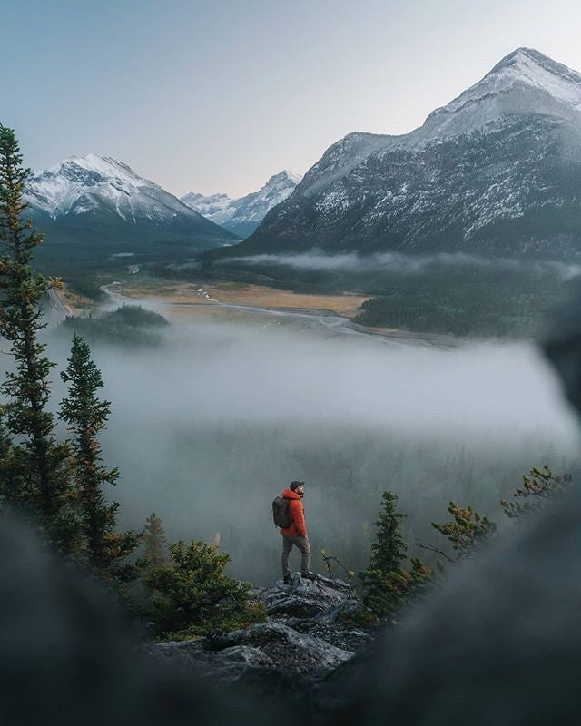 A man with a waterproof camera backpack standing on a rock looking out at fog and trees