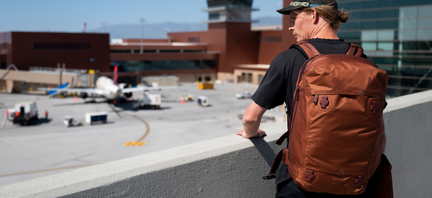 A man in a black shirt and hat stares across an airport with his Arris Pack on his back.
