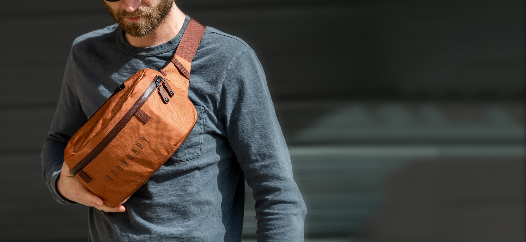 A man in a gray-blue shirt and sunglasses wearing his Boundary Supply Sling pack.