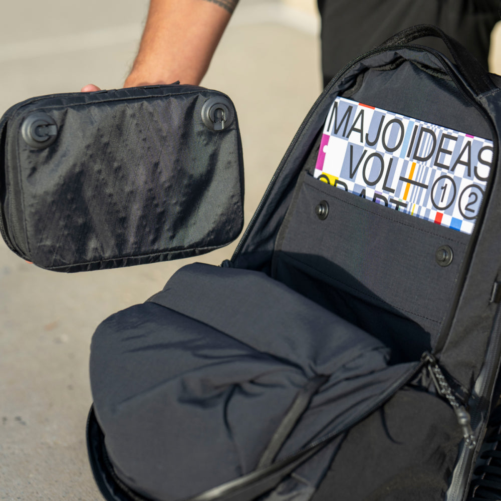Black backpack with a visible book titled 'MAJO IDEAS VOL. 2' on a concrete surface.