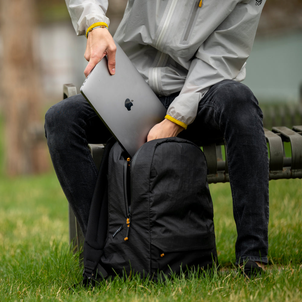 Person sitting on a bench holding a laptop with a backpack nearby, outdoors.